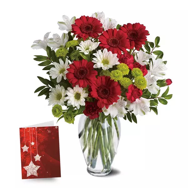 A bouquet of red, white, and green flowers in a clear vase next to a red greeting card.