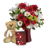 A teddy bear alongside a vibrant bouquet in a red vase with a love-themed card.