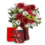 A bouquet of red and white flowers in a vase with a box of chocolates and a card with hearts.