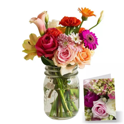 A bouquet of mixed flowers including roses and daisies in a mason jar, with a floral card beside it.
